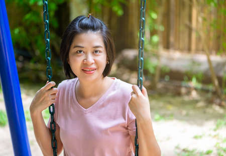 Portrait of smiling Asian woman in children's playground, Asian middle aged woman sitting on outdoors swing playground, smiling face, black short hair, eyes looking at camera.のeditorial素材