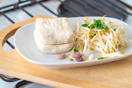 Organic bean sprouts, tofu, garlics and scallions in white plate which on wooden cutting board in...
