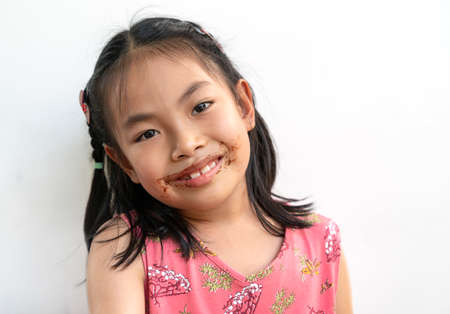 Portrait beautiful child girl with chocolate around her mouth, big smile on cute face, black hair, wearing beautiful pink dress. Beautiful Asian child girl on white background.の写真素材