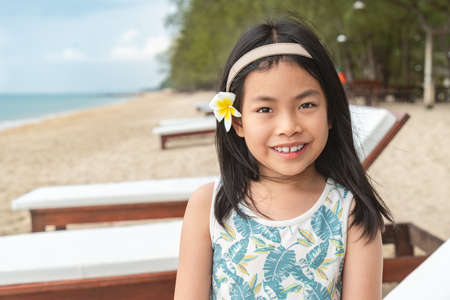 Portrait of Asian child girl on a beach of Thailand, black long hair with white flower at ear as decoration, sit on sunbed of the resort, eyes looking at camera, smiling. Background sea and sky.の写真素材