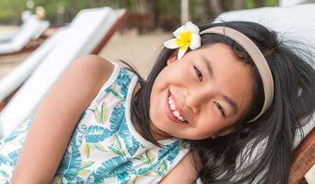 Portrait of Asian child girl leaning on sunbed on a beach of Thailand, black long hair with white flower at ear as decoration, smiling face, eyes looking at camera.の写真素材
