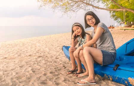 Beautiful Asian mother and child sit on kayak boat, pose on the sandy beach, blurred background of sea and sky, perspective view, warm tone image with blank space for copy and design.の写真素材