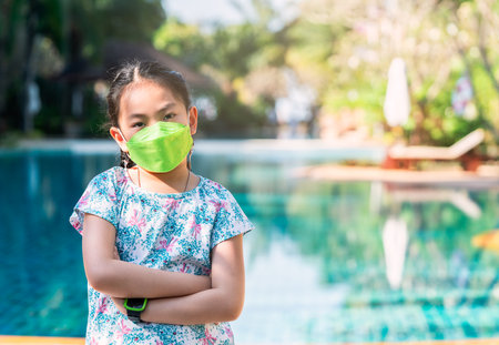 Portrait Asian child girl wearing a face mask, arms on the chest and standing beside swimming pool at a resort, concept for traveling in the era of virus in Thailand. Day light image in a morning.の写真素材