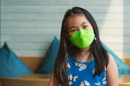 Portrait Asian little girl wears a face mask in a restaurant, black long hair child girl, natural light from the window.の写真素材