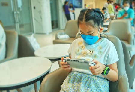 Portrait Asian child girl is playing a portable game handheld console while waiting for a doctor at a hospital, wearing medical face mask, blurred background of hospital atmosphere.の写真素材