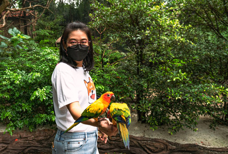 Portrait of Asian middle-aged woman in a zoo, holding cute Sun conure birds, wearing a black color...