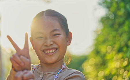 Happy Asian child girl with sunset light, light flare from behind, happy face, show sign of happiness with two fingers gesture, healthy child girl at 9 to 10 years old, bokeh background from tree.の写真素材