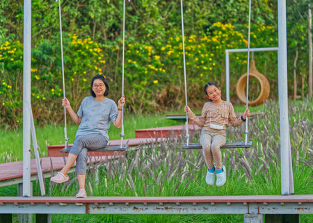 Asian middle-aged mother sitting on a wooden swing in the garden with a little child girl or...