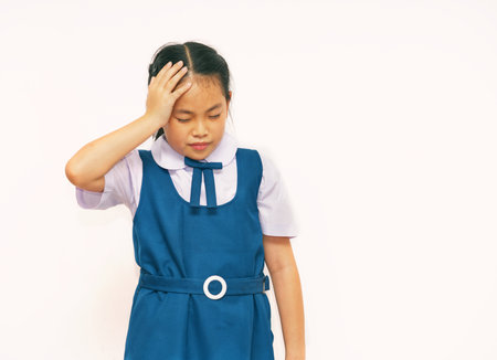 Asian girl in blue school uniform, hand on head, showing discomfort or headache, eyes slightly closed, plain light background, expression of pain or fatigue. Ideal for education or health topicsの写真素材