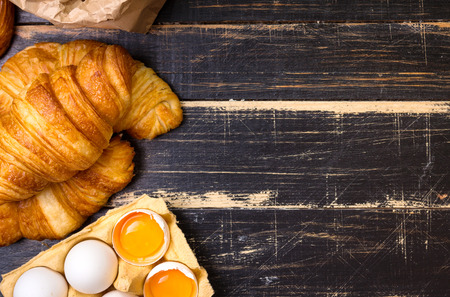 Freshly baked croissants with flour, wooden spoon, eggs and egg yolks in a carton tray on the dark...