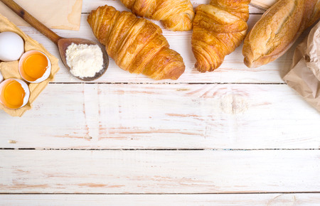 Freshly baked croissants and baguette with flour, wooden spoon, piece of paper, eggs and egg yolks in a carton tray on the white wooden table. Bakingpastry background. Free space for textの写真素材