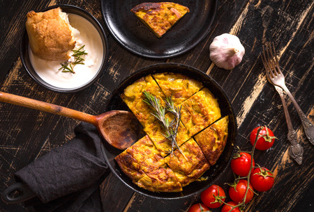 Tortilla de patatas in a pan with garlic sauce aioli and fresh tomatoes cherry. Traditional spanish dish. Omelette with eggs, potatoes and onion. Rustic black background. Top viewの写真素材