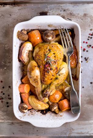 Whole baked juicy chicken with golden crispy skin. Roasted chicken in baking dish with apples, carrot, mushrooms, rosemary, garlic, pepper on rustic iron oven-tray. Overhead. Dinner. Cooked poultryの写真素材