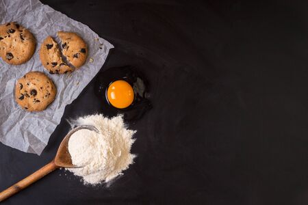 Rustic wooden spoon with a flour, a raw egg, a baking paper and cookies on a black chalkboard...