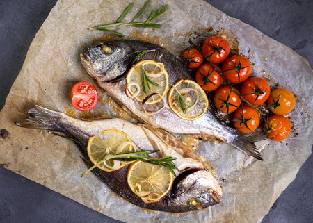 Tasty baked whole fish on baking paper. Baked sea bream with lemon, onion, herbs, cherry tomatoes, spices on dark rustic background. Grilled delicious fish. Diet and healthy food. Top viewの写真素材