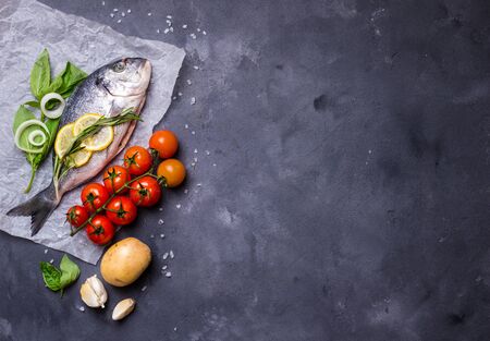 Raw fish with fresh ingredients ready to cook. Fish, lemon, herbs, potato, tomatoes. Ingredients for cooking on dark rustic background. Space for text. Diet and healthy food. Fish background. Top viewの写真素材