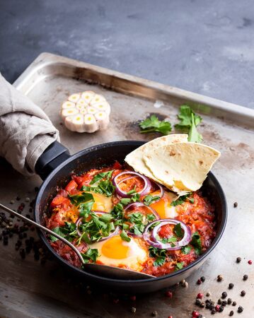 Shakshuka with pita bread in a pan. Middle eastern traditional dish. Fried eggs with tomatoes, bell pepper, vegetables and herbs. Shakshouka on a table. Sunny side up eggs. Homemade. Selective focusの写真素材