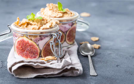 Homemade healthy yoghurt in glass pot with cereals, figs, mint on rustic concrete background. Healthy morning breakfast. Freshly made yoghurt with oat granola and fruit. Yoghurt in jar with muesliの写真素材