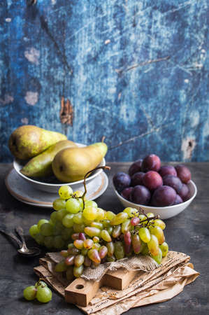 Fresh ripe grapes on wooden cutting board, pears and plums in bowls, wooden rustic background. Fresh juicy organic fruits. Close up. Organic natural fruits. Autumn/fall setting. Harvest/crop/gardenの写真素材