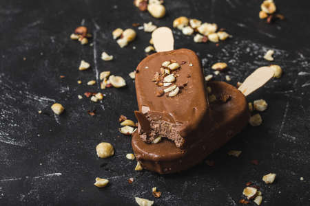 Milk chocolate ice cream with hazelnuts. Close-up. Ice cream covered with chocolate, sticks, black stone background. Selective focus. Chocolate ice cream bars, nuts. Dessert. Bitten ice creamの写真素材