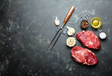 Raw marbled meat steak Ribeye on black rustic stone background with fork, seasonings and olive oil for cooking, roasting, grill or BBQ party with space for text, top view. Meat restaurant conceptの写真素材