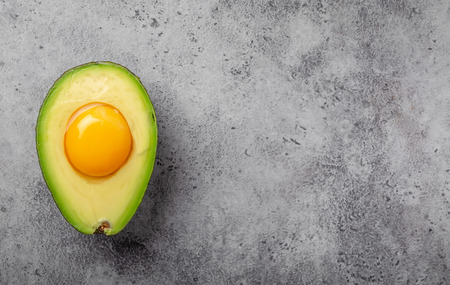 Foods rich in healthy fats for balanced nutrition: raw egg yolk in fresh cut half avocado on gray...