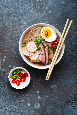 Tasty Japanese noodle soup ramen in white ceramic bowl with meat broth, sliced pork, narutomaki, egg with yolk on rustic stone background. Traditional dish of Japan, top view, close-up, conceptの写真素材