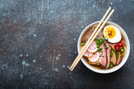 Tasty Japanese noodle soup ramen in white ceramic bowl, meat broth, sliced pork, narutomaki, egg with yolk on rustic stone background. Traditional dish of Japan, top view, close-up. Space for textの写真素材