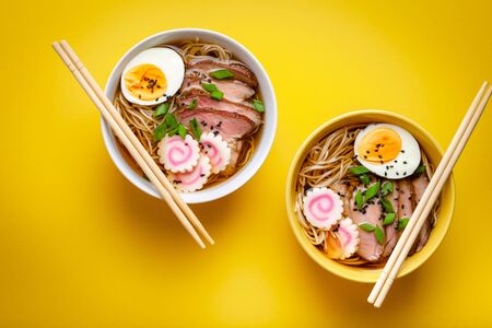 Two bowls of Japanese noodle soup ramen with meat broth, sliced pork, narutomaki, egg with yolk on pastel yellow background. Traditional dish of Japan, top view, close-up, conceptの写真素材