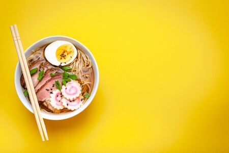 Tasty Japanese noodle soup ramen in white ceramic bowl, meat broth, sliced pork, narutomaki, egg with yolk on pastel yellow background. Traditional dish of Japan, top view, close-up, space for textの写真素材