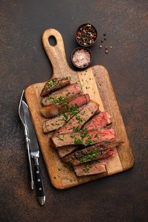Grilled or fried prime marbled meat steak Ribeye sliced and served on wooden cutting board with cutlery. Top view of juicy cooked beef steak cut in slices on rustic brown concrete backgroundの写真素材