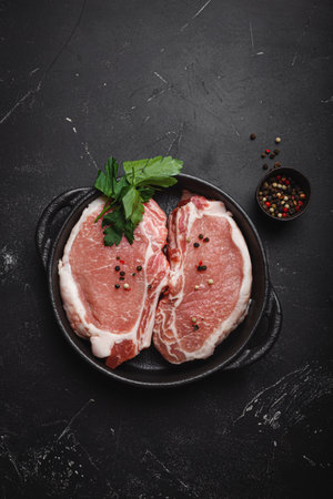 Cut raw meat pork steaks with seasonings in black cast iron pan on dark rustic stone background top view, ready for roasting. Pork loin chops cookingの写真素材