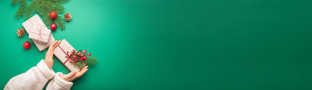 Christmas or New Year celebration green paper festive background with female hands holding wrapped present box, decoration fir tree, cones, berries, sparkly red balls. Space for text.の写真素材