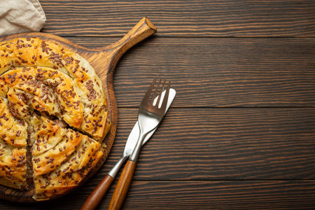 Burek made of filo dough with filling on cutting board, dark brown wooden rustic background top view. Traditional savoury spiral pie of Balkans, Middle East and Central Asia. Space for textの写真素材