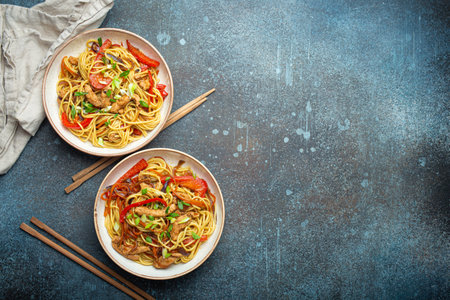 Two bowls with Chow Mein or Lo Mein, traditional Chinese stir fry noodles with meat and vegetables, served with chopsticks top view on rustic blue concrete background, space for textの写真素材