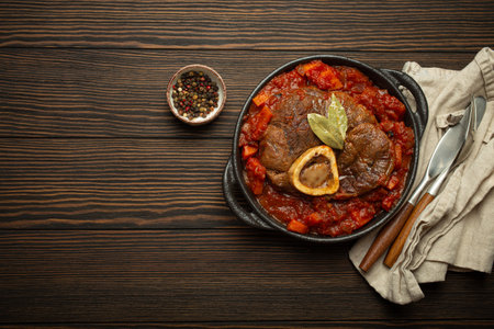 Traditional Italian dish Ossobuco all Milanese made with cut veal shank meat with vegetable tomato sauce served in black casserole pan top view on rustic brown wooden background, copy spaceの写真素材