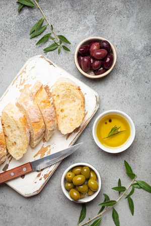 Sliced fresh ciabatta on cutting kitchen board, green and brown olives, olive oil with rosemary, olive tree branches on gray concrete stone rustic background top view copy spaceの写真素材