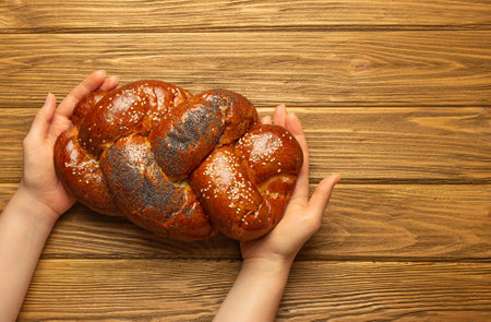 Female hands holding freshly baked Challah bread covered with poppy and sesame seeds, top view on rustic wooden background, traditional festive Jewish cuisineの写真素材