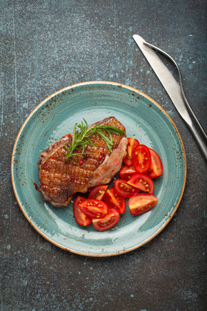 Delicious roasted duck breast fillet with golden crispy skin, with pepper and rosemary, top view on ceramic blue plate served with cherry tomatoes salad, rustic concrete backgroundの写真素材