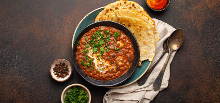 Traditional Indian Punjabi dish Dal makhani with lentils and beans in black bowl served with naan flat bread, fresh cilantro and two spoons on brown concrete rustic table top view. Space for textの写真素材