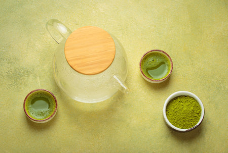 Steaming glass teapot and matcha green tea powder with two cups of prepared tea on green background top viewの写真素材