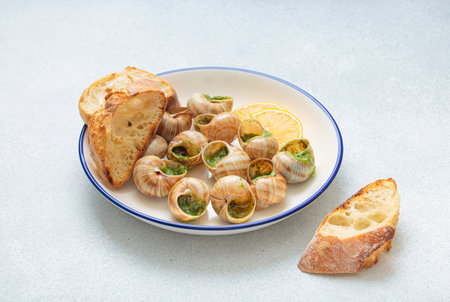 Escargots de bourgogne snails with garlic butter, parsley and lemon wedges on white plate on light backgroundの写真素材