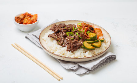 Korean beef bulgogi bowl dish with rice, roasted meat, kimchi and marinated cucumber on white background with chopsticksの写真素材