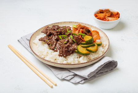 Korean beef bulgogi bowl dish with rice, roasted meat, kimchi and marinated cucumber on white background with chopsticksの写真素材