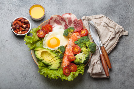 Balanced ketogenic diet meal with salmon, avocado, egg, bacon and vegetables on plate with cutlery top view on grey backgroundの写真素材