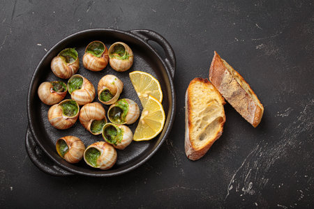 Snails escargots with garlic butter and lemon, served in a black pan with toasted bread baguette slices on black rustic backgroundの写真素材