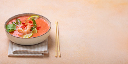 Traditional Thai soup tom yum soup with shrimp, lime, and herbs in bowl with chopsticks on light...