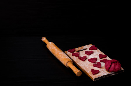 Raw red heart-shaped cookies and dough on wooden board with flour, wooden rolling pin. Black background. Preparing for Valentine's Dayの写真素材