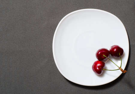 Three cherry on the edge of an empty plate on the texture gray background, space for text, top viewの写真素材