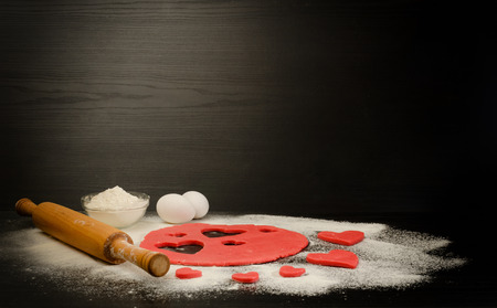 Red dough, cut out hearts, flour, eggs and rolling pin on a black background, space for textの写真素材